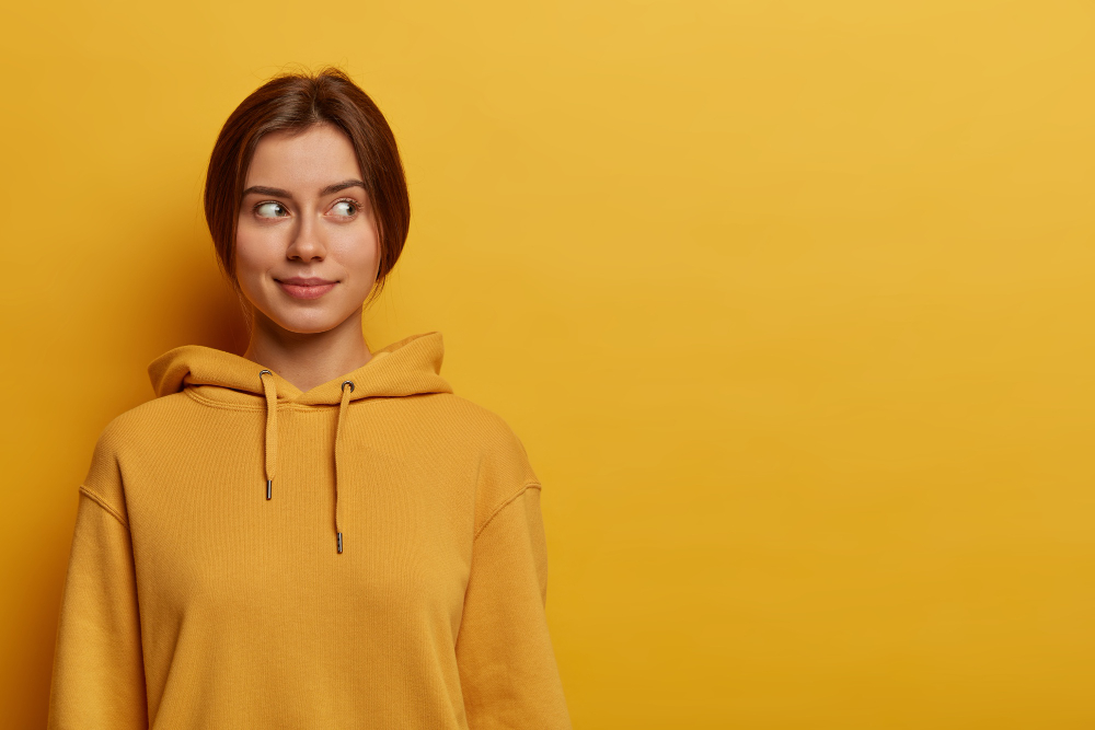 shot-of-pretty-teenage-girl-looks-thoughtfully-aside-has-dark-combed-hair-wears-casual-sweatshirt-poses-against-yellow-wall-thinks-about-going-on-picnic-with-friends-at-weekend.jpg