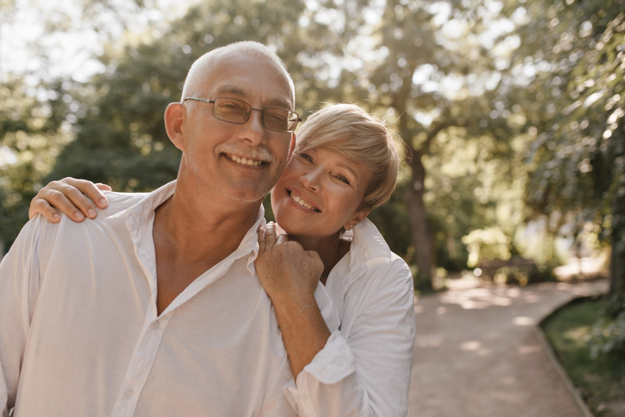 smiling-old-man-with-grey-hair-and-mustache-in-glasses-and-light-shirt-hugging-with-blonde-woman-in-white-clothes-in-park.jpg