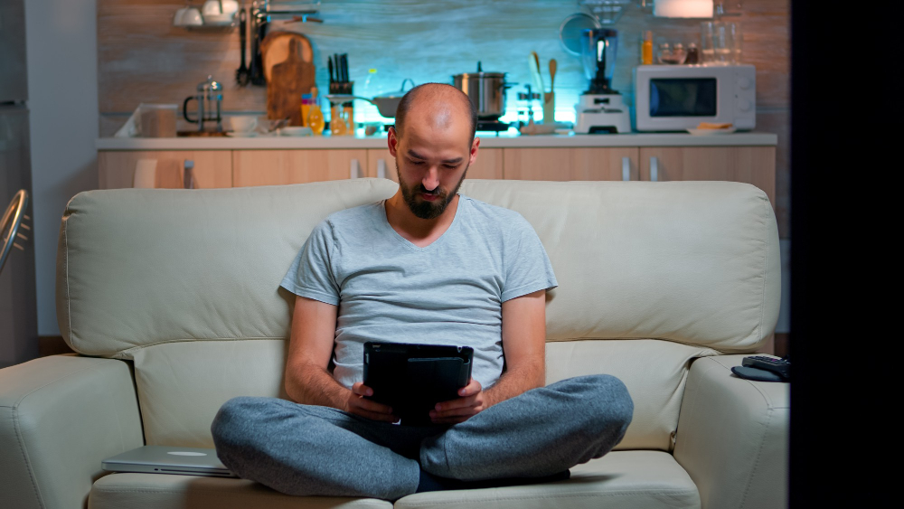 tired-man-sitting-alone-on-couch-while-browsing-on-internet-using-tablet-computer-with-modern-technology-wireless-caucasian-male-in-pajamas-relaxing-in-front-of-televion-late-at-night-in-kitchen.jpg